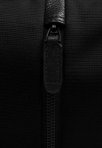 Die schwarze Nylon-Tasche verfügt über eine strukturierte Oberfläche, einen robusten Reißverschluss mit einem Lederzieher und verstärkte Nähte entlang der Kanten.