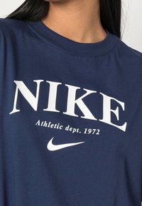 Tmavě modré bavlněné tričko s velkým bílým nápisem "NIKE", menším nápisem "Athletic dept. 1972" pod ním a logem Nike swoosh. Krátké rukávy.