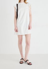 Valkoinen lyhyt mekko, jossa on röyhelöhihoja, teksturoitu kangas ja rypistyskohta bodicessa. Yhdistetty mustiin korsettikorkoihin.