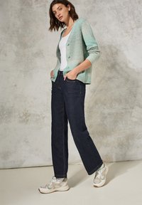 Stickad cardigan i ljus blågrön med tre knappar, i kombination med mörka raka jeans och sneakers med en beige och grön design.