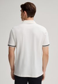 Polo blanc en tissu texturé, avec des accents rayés noirs et blancs sur le col et les manches. Étiquette visible en bas.