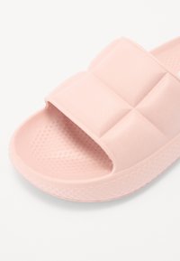 Pink slip-on sandals made of soft material, featuring a textured footbed and a padded strap with a quilted design. Circular perforations are visible.