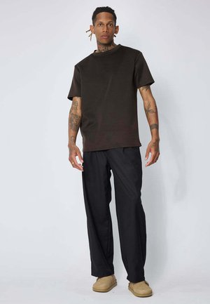 Jeune homme tatoué portant un t-shirt marron foncé, un pantalon noir et des chaussures beige, debout devant un fond clair uni.