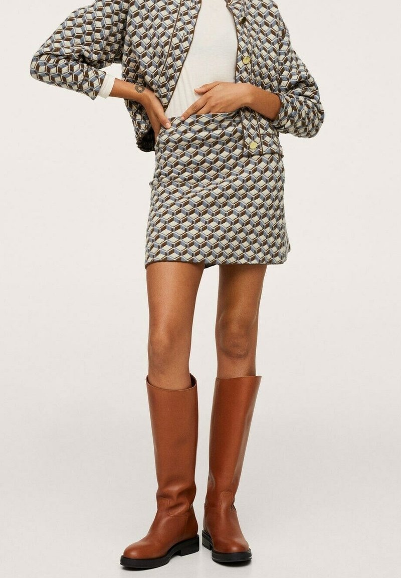 Femme portant une veste à motif géométrique et une minijupe avec un haut blanc et des bottes en cuir marron hauteur genoux, posant les mains sur les hanches.