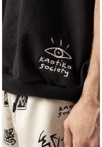 Czarna bluza z haftowanym logo "kaotiko society" i wzorem oka, w połączeniu z białymi spodniami z czarnymi abstrakcyjnymi wzorami.