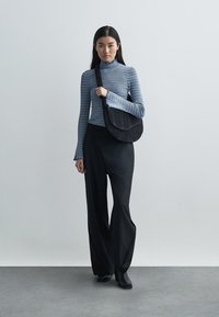 Sac à bandoulière noir de forme arrondie et texture rayée. Porté par une personne en pull col roulé à côtes bleu et pantalon large noir.