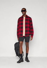 Rød og svart rutete flanellskjorte, beige t-skjorte, svarte shorts, solbriller, svart ryggsekk, svarte joggesko, atletiske sokker.