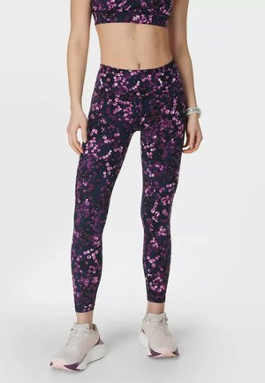 Vrouw draagt donkerpaarse en roze legging met bloemenpatroon, lichtroze sneakers en een fitness tracker om de pols.