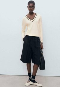 Maglione beige a scollo a V con rifiniture nere, abbinato a pantaloni doppiopetto neri. Gli accessori includono una borsa nera e scarpe con lacci color crema.