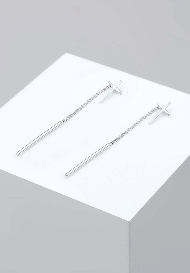 Boucles d'oreilles en argent présentant un design de croix minimaliste sur une chaîne, avec des tiges allongées pendues en dessous, exposées sur un piédestal blanc.