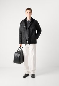 Black waterproof jacket with two chest pockets, layered over a patterned sweater. Model holds a black leather bag, wears white pants and shoes.