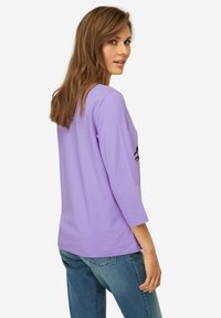 Camicia a maniche lunghe lavanda con scollatura rotonda e maniche a tre quarti, con ricami neri sui lati. Tessuto morbido e liscio.