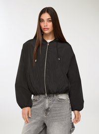 Giacca bomber leggera nera con cappuccio, zip argentata e polsini elasticizzati; abbinata a jeans in denim grigi a vita alta.