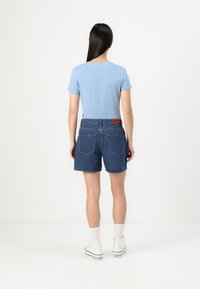 Light blue short-sleeve t-shirt paired with dark denim shorts. Shorts feature two back pockets and a branded brown leather patch. White sneakers visible.