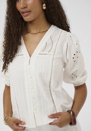 Bluză albă din material ușor, cu modele decupate elaborate, mâneci bufante și nasturi în față cu margine decorativă.