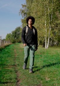 Oseba hodi po travnati poti, oblečena v črno jakno, zelene hlače in sivo obutev, z nahrbtnikom in afro frizuro.