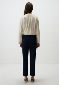 Blouse en crème avec détail froncé au dos, associée à un pantalon noir marine cintré et des sandales à talons nude, vue de dos.