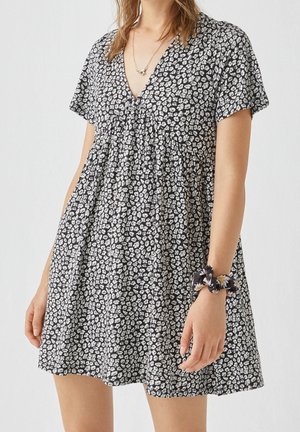 Femme portant une courte robe noire à motif floral blanc, décolleté en V, manches courtes et chouchou assorti au poignet.