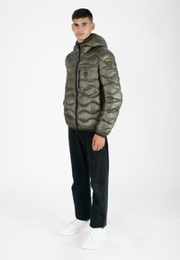 Olivegrüner Daunenjacke mit Kapuze, Reißverschluss vorne und strukturiertem, gesteppter Design. Kombiniert mit schwarzen Hosen und weißen Sneakers.