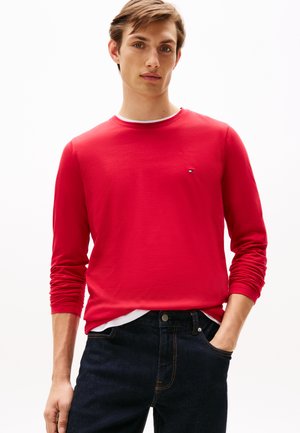 Jonge man die een rood langemouwen shirt over een witte t-shirt draagt, met donkere spijkerbroeken en één hand in zijn zak, kijkend naar voren.