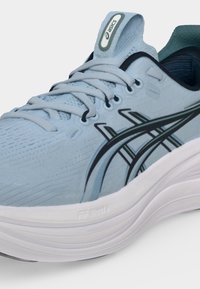 Ανοιχτό μπλε αθλητικό παπούτσι τρεξίματος Asics με λευκή χοντρή σόλα, ανοιχτογκρι κορδόνια, μαύρες και μπλε πλαϊνές ρίγες και επισημασμένη ετικέτα στη γλώσσα.