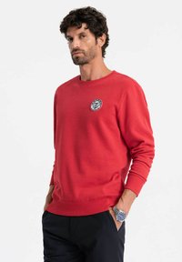 Sudadera roja con cuello redondo y mangas largas. Presenta un parche de logo redondo en blanco y negro en el pecho. Textura suave, ajuste relajado.