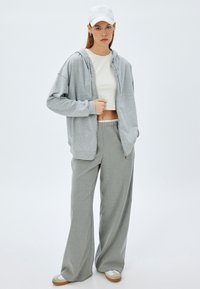 Sudadera con cremallera de color gris claro y un corte relajado, llevada sobre un top corto blanco. Combinada con pantalones de pierna ancha de rayas grises y zapatos blancos.