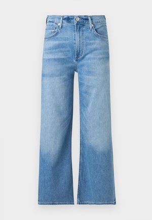 Naine, kes kannab musta varrukateta topi, hele sinist laiade säärtega teksapükse musta vööga ja musti kontsaga sandaale, seisab ühe käega taskus.