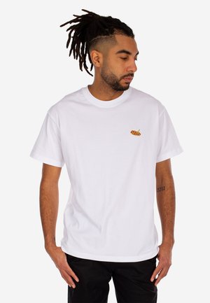 Mann mit Dreadlocks, der ein weißes T-Shirt mit einem kleinen Spieß-Essen-Design auf der Brust trägt und nach unten zu seiner linken Seite schaut.