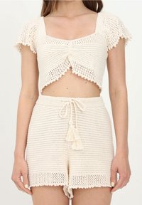 Femme portant un crop top en crochet de couleur crème avec manches courtes et un short taille haute assorti avec cordon et pompons.
