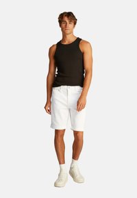 Camiseta sin mangas negra combinada con pantalones cortos de denim blancos, con dobladillos enrollados. Modelo con zapatillas blancas y calcetines blancos de longitud media.