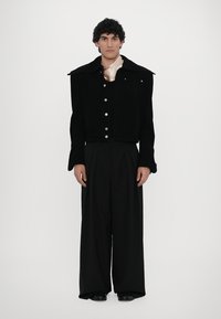 Black corduroy jacket with a wide collar, silver buttons, and an embroidered detail, paired with wide-legged black trousers. Smooth silk neck scarf.