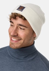 White ribbed beanie with a square patch displaying "JEANS CODE" in orange and navy. Worn with a dark blue turtleneck sweater.