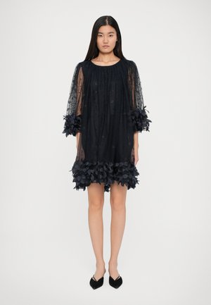 Woman wearing a black sheer dress with floral 3D embroidery on sleeves and hem, paired with black pointed-toe flats, standing on white background.
