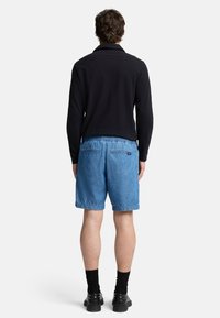 Polo shirt nero a maniche lunghe, pantaloni corti in denim blu con vita elastica, calze scure e scarpe nere, vista da dietro.
