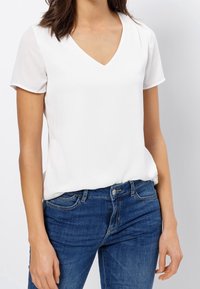 Weißes kurzärmliges V-Ausschnitt-Shirt, glatte Textur, kombiniert mit blauen Denim-Jeans, die klassische Taschen und ein tailliertes Design aufweisen.