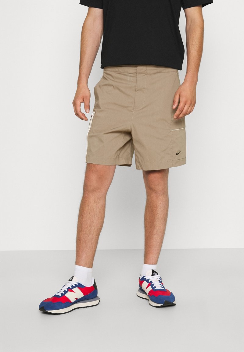Shorts de sport beiges en tissu léger avec une poche latérale zippée, associés à des baskets multicolores aux accents rouges, blancs et bleus.