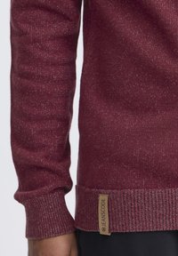 Burgunderfarbener Strickpullover mit gerippten Bündchen und Saum, der ein strukturiertes Muster aufweist und ein hellbraunes Stoffetikett mit der Aufschrift "JEANSCODE" an der unteren Seite trägt.