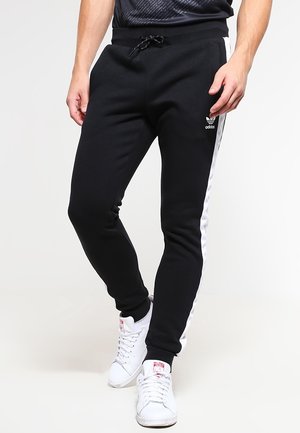 Mand iført sorte Adidas joggingbukser med hvide siderstriber og hvide sneakers, stående med hænderne afslappet ned langs siderne.