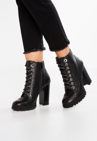 Botas de cuero negras hasta el tobillo con un tacón alto y cuadrado, cordones en la parte delantera con ojales plateados, cierre con cremallera y suela de goma texturizada.