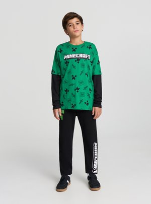Maglietta verde a tema Minecraft con maniche lunghe nere, caratterizzata da disegni pixelati; abbinata a pantaloni neri e scarpe da ginnastica nere.
