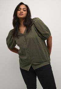 Blouse verte à manches bouffantes avec décolleté en V et boutons sur le devant. Fabriquée dans un tissu léger et texturé. Associée à un pantalon sombre.