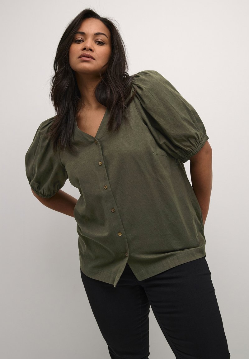 Blouse verte à manches bouffantes avec décolleté en V et boutons sur le devant. Fabriquée dans un tissu léger et texturé. Associée à un pantalon sombre.