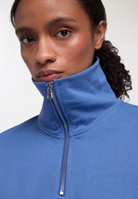 Felpa blu con collo alto realizzata in tessuto morbido, caratterizzata da una chiusura con zip a un quarto e una superficie liscia.