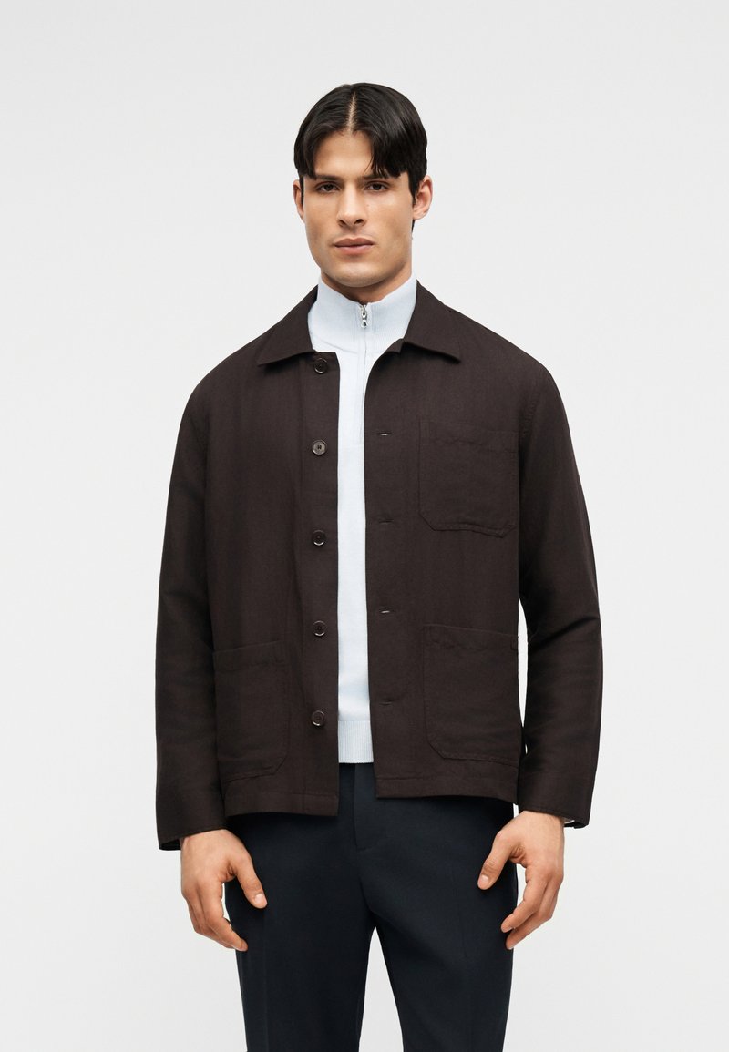 Muž v tmavě hnědém saka na knoflíky přes bílý top na zip a tmavé kalhoty, stojící před jednoduchým světlým pozadím.