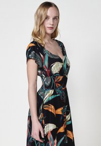 Vestido negro con estampados vibrantes de hojas tropicales en verde, naranja y amarillo. Presenta un escote en V y una cintura fruncida para una silueta ajustada.