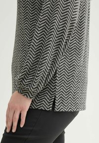 Houndstooth-patroon top in grijs en zwarte stof, met een zij-split en losse mouwen. Draagt over zwarte onderkleding, wat de textuur benadrukt.