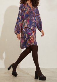 Modré šaty s dlouhými rukávy, pokryté květinovými a abstraktními vzory. Nosené s černými lodičkami na vysokém podpatku a tmavými punčocháči.