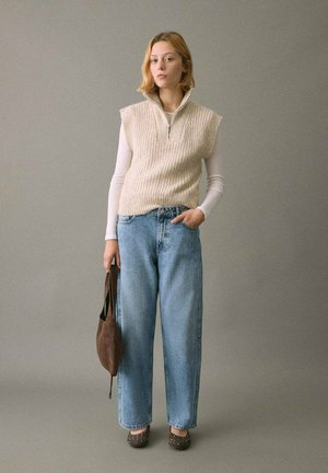 Chaleco de punto beige con cuello de cremallera sobre una camisa blanca de manga larga, combinado con pantalones vaqueros de pierna ancha en azul claro y zapatos marrones con tachuelas.