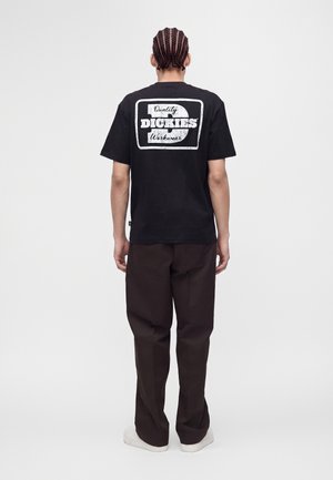 Uomo con i capelli intrecciati, visto di spalle, indossa una maglietta nera Dickies, pantaloni marrone scuro e scarpe bianche su sfondo uniforme.